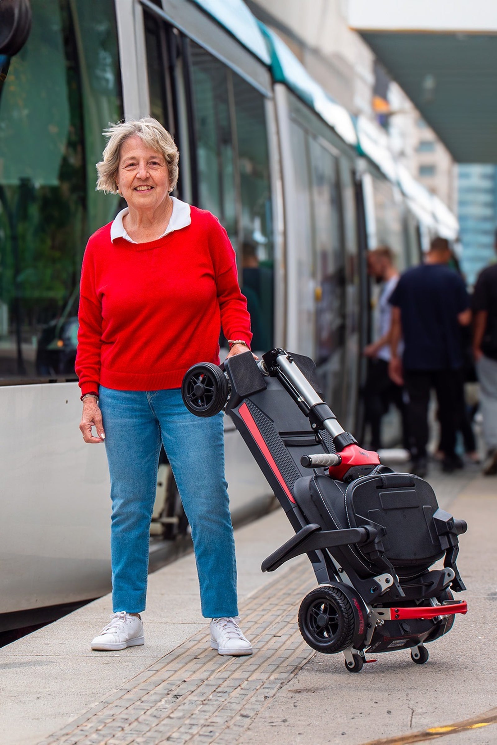 An elderly woman in a red sweater stands on a train platform next to the folded Muvon One scooter. The scooter is tilted upright in "trolley mode," resting on its small transport wheels while she holds the handle, illustrating how easily it can be wheeled like a suitcase near public transportation.