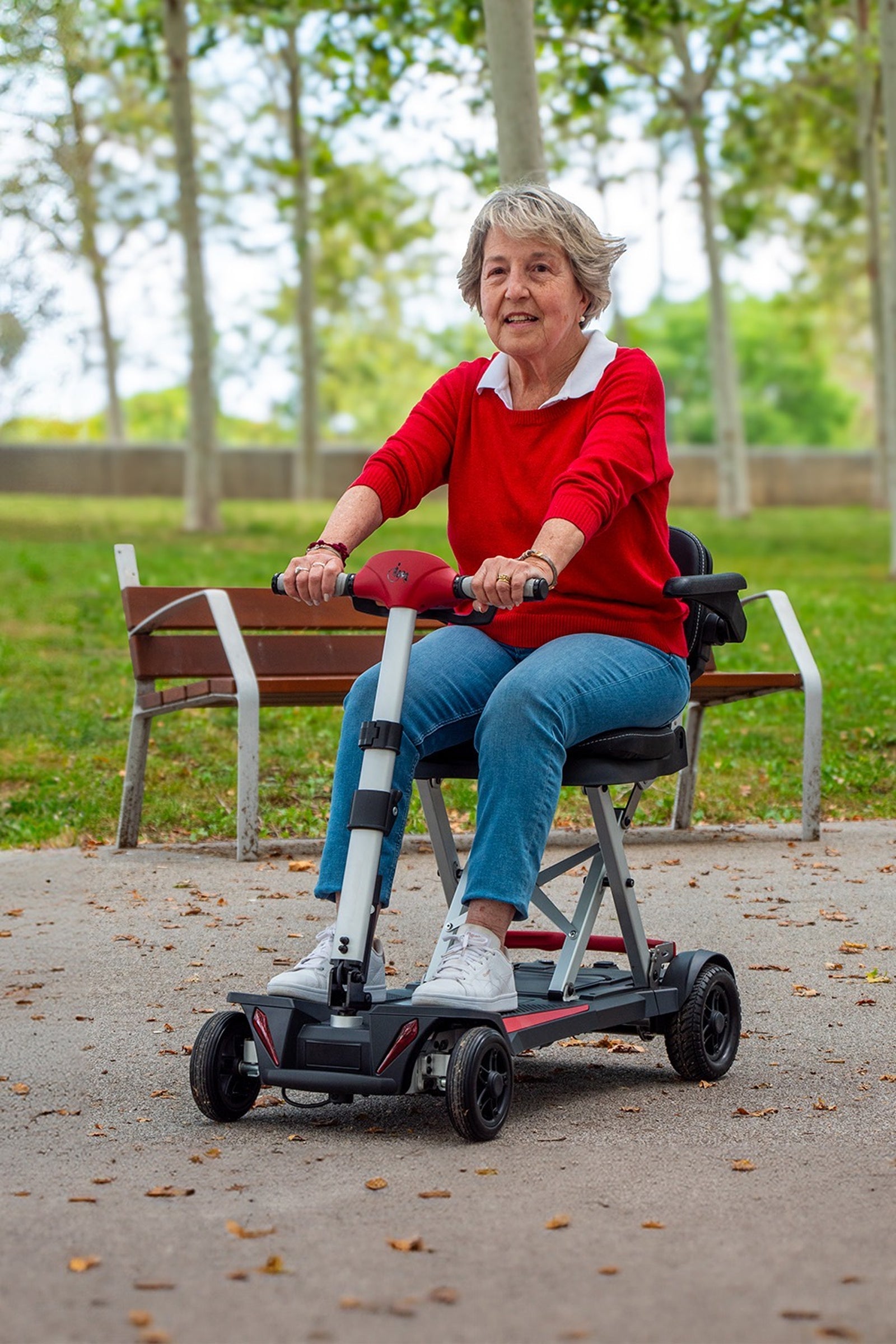 The woman riding the Muvon One scooter along a smooth park path lined with trees and benches. The front-facing angle shows her comfortably gripping the ergonomic red handlebars. The scooter’s compact four-wheel footprint and high ground clearance are visible as it maneuvers through a natural, recreational setting.