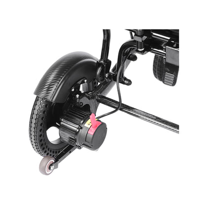 An angled close-up view of the rear motor and drivetrain assembly of the Harvey Carbon Power Wheelchair. It shows a large rear drive wheel with a unique textured-tread pattern, partial carbon fiber fender with a detailed weave, and the compact electric hub motor. A bright red manual freewheel lever is prominent on the motor, and a small anti-tip caster wheel is positioned further rearward. 