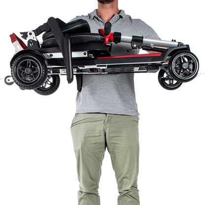 A person holding the Muvon One mobility scooter horizontally with both hands to demonstrate its lightweight design. The scooter is fully folded into a compact, rectangular shape with the red tiller tucked against the black seat and the four wheels visible at the corners.