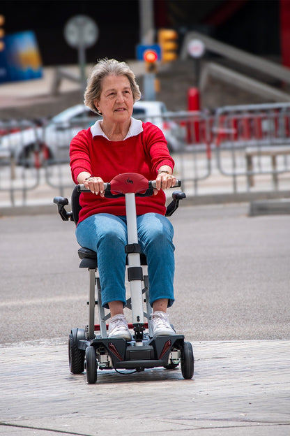 Front view of an elderly woman riding the Muvon One mobility scooter across a paved outdoor plaza. The scooter is fully unfolded, showing its slim white steering column, red dashboard, and compact four-wheel base. The woman is smiling, with her hands on the handlebars, demonstrating the scooter’s stability and ease of use in an urban environment.