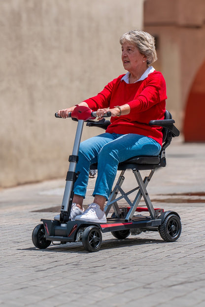 Side view of an elderly woman operating the Muvon One scooter on a paved cobblestone street. The image highlights the scooter's X-frame seat support and its low-profile floorboard. The woman is looking ahead, emphasizing the scooter’s suitability for outdoor travel and daily errands.