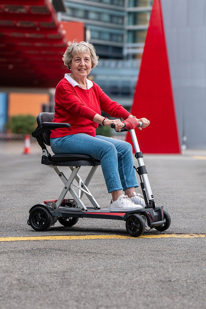 A wide-angle shot of the woman riding the Muvon One scooter through a modern city area with large architectural structures in the background. The scooter is shown in a three-quarter profile, displaying the black padded seat with armrests and the red accents on the base and tiller, framing it as a stylish urban mobility solution.