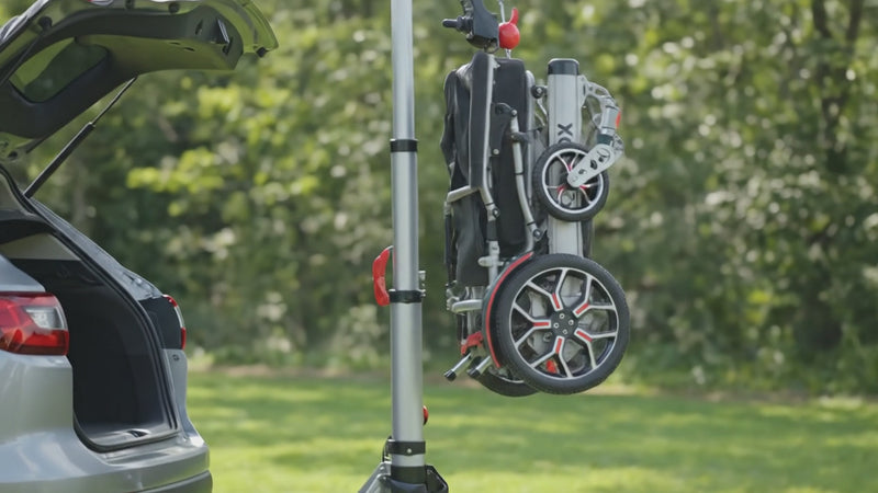 Video set in a lush green outdoor environment during soft daylight. A modern silver vehicle is parked with its trunk open. Next to the vehicle stands the Muvon Mobility portable lift. It operates smoothly and quietly, showcasing precise engineering and stability. Attached securely to the lift mechanism is the folded Muvon Equinox power wheelchair.

The wheelchair is being lifted vertically from ground level in a controlled, steady motion. The movement is fluid and effortless, empha