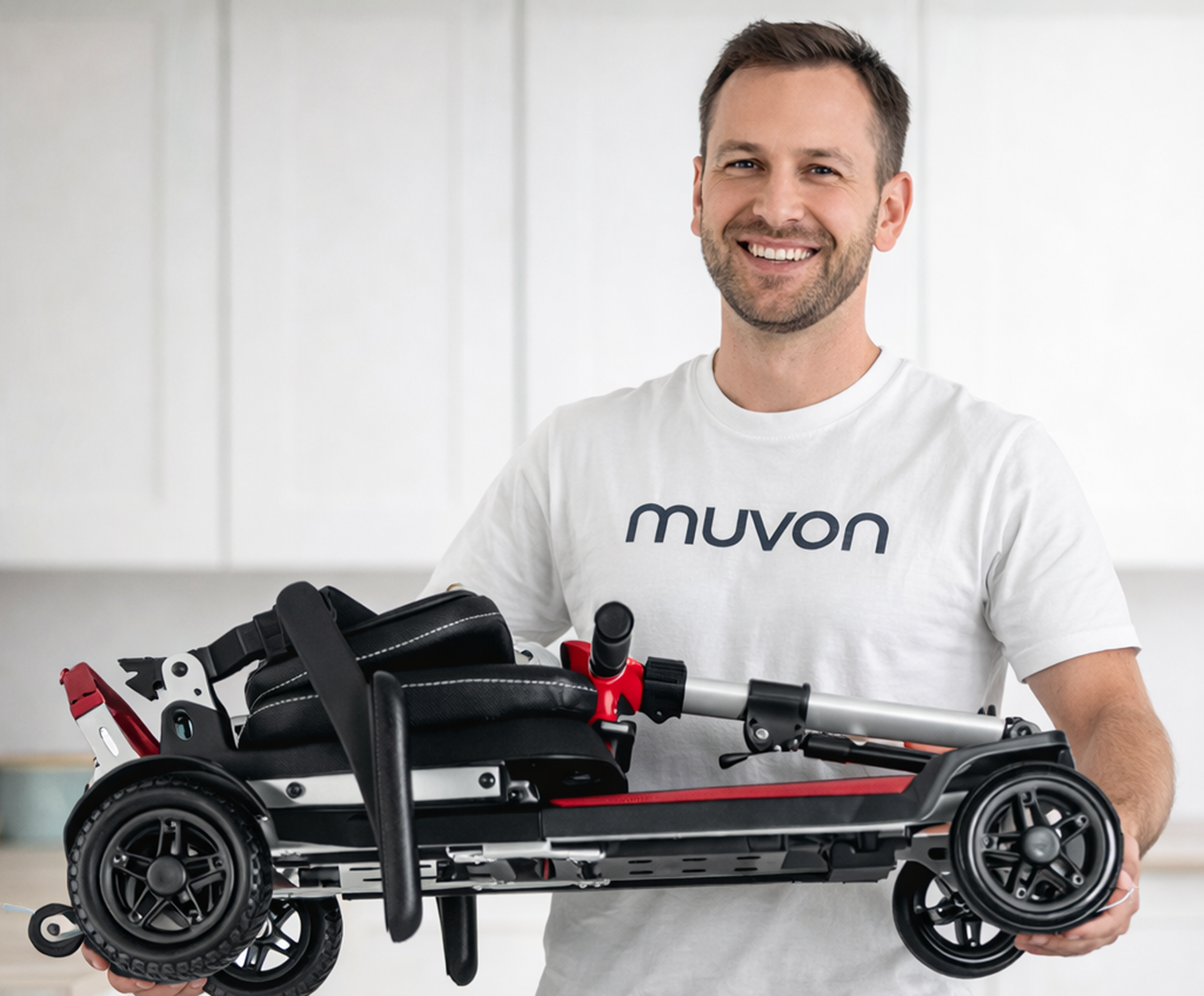 A smiling Muvon Mobility service technician wearing a white t-shirt with the "muvon" logo holds a Muvon One mobility scooter that has been fully folded into a compact, horizontal position. The scooter features a red and black frame with silver accents. The man is standing in a brightly lit indoor space with a blurred white kitchen background, demonstrating the scooter's portability and lightweight design.